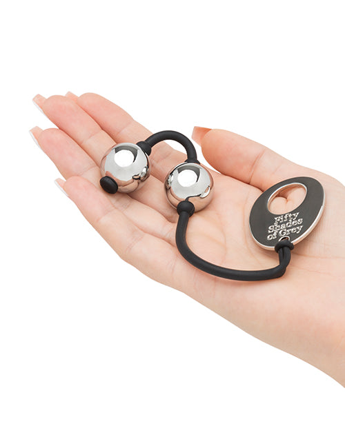 The Fifty Shades of Grey Inner Goddess Kegel Balls sitting inside of the flattened palm. The balls look particularly small and slim in the hands. The end of the kegel balls string has a large metal tag that has the "Fifty Shades of Grey" logo written on it. | Kinkly Shop