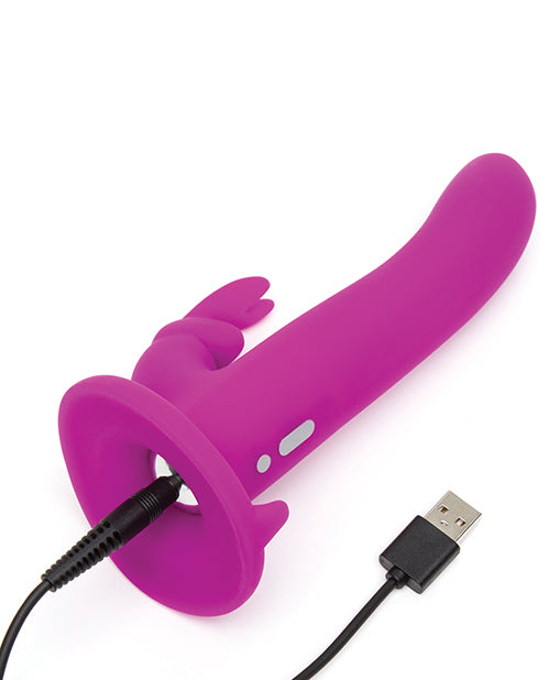 The rabbit vibrator of the Happy Rabbit Strap On Harness Set plugged into its charger. The charging port is underneath the toy itself, hidden at the base of the flare underneath the toy. | Kinkly Shop