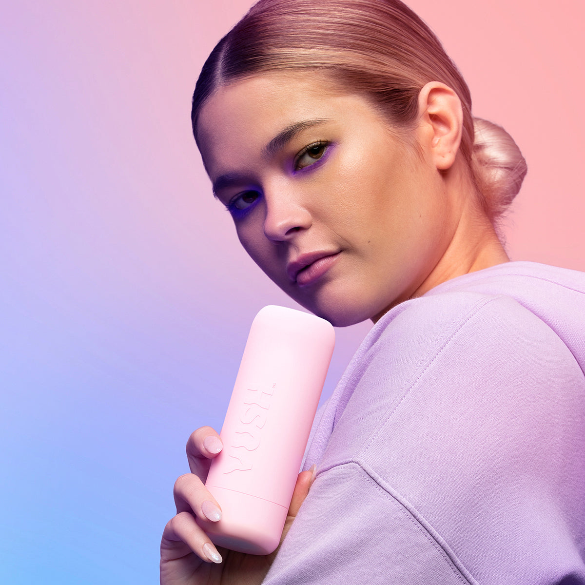 A person looks into the camera while holding the VUSH Glow travel case. The case looks like a relatively discreet cylinder in pink that only has the text "VUSH" written on it. | Kinkly