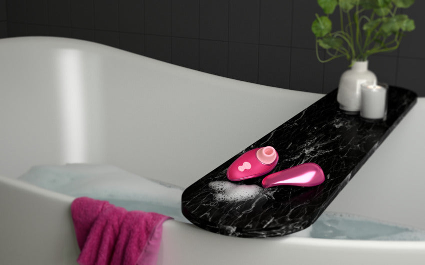 The Womanizer x Iggy Azalea Liberty 2 is resting on the edge of a bathtub with the lid lying next to it. It looks very small and only slightly bigger than the little candle that's shown in the background. | Kinkly Shop