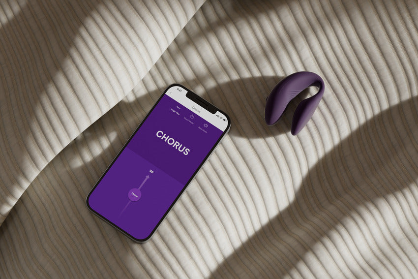 The We-Vibe Chorus lays out on a textured bedspread next to a cell phone. The cell phone is open to the We-Vibe Connect app, showcasing the slider control that would allow the phone to control the vibrator. | Kinkly Shop