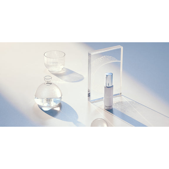 A bottle of the LELO Antibacterial (Toy) Cleaning Spray 2 fl oz sits out on a white surface with a strong sunbeam shining into the image from the corner. There are other white, see-through objects like glasses, cups, and blocks. All of the items cast soft, long shadows in the sunbeam. The toy cleaner bottle does as well. | Kinkly Shop