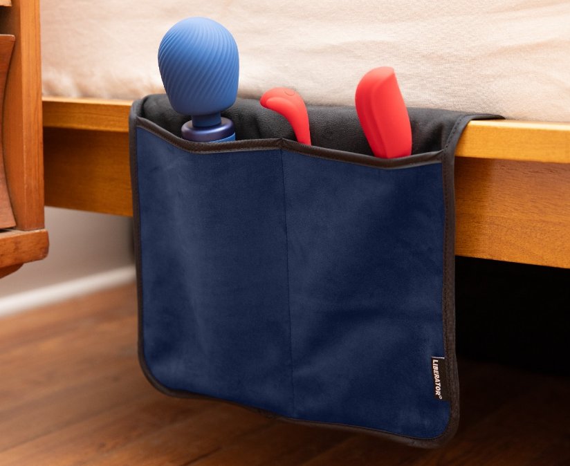 The Liberator Fascinator Sex Toy Caddy tucked underneath a mattress and displayed next to a bed. Two sex toys, including the Fun Factory VIM, are shown peeking out from the tops of the pockets. | Kinkly Shop