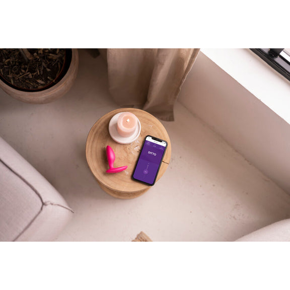 The We-Vibe Ditto+ in Cosmic Pink sitting on a wooden end table. A lit candle and a cell phone, connected to and controlling the We-Vibe Ditto+, are shown next to it. | Kinkly Shop