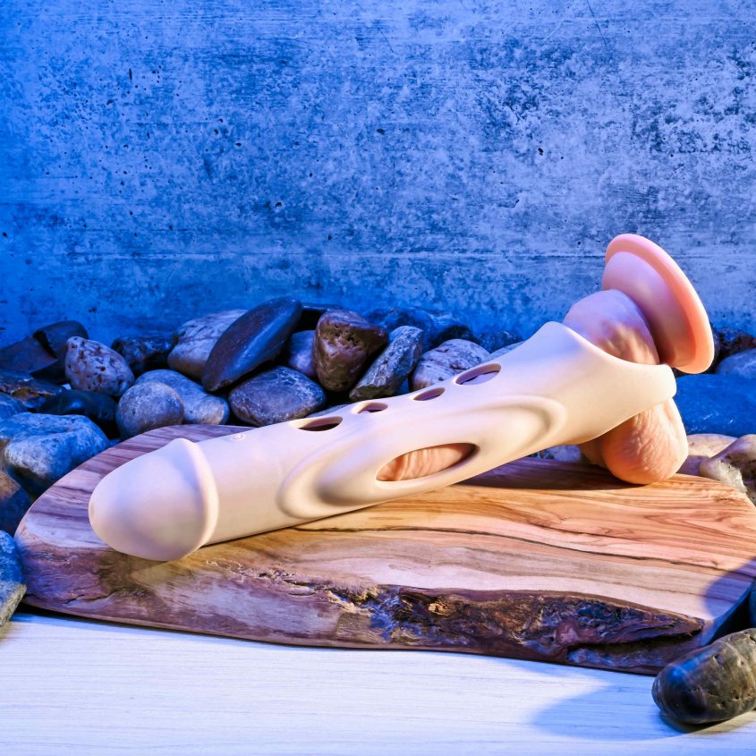 The Evolved Big Shaft Extender in Cream shown overtop of a dildo in front of a textured background. The peek-a-boo holes make it easy to see the dildo underneath the extender itself. | Kinkly Shop