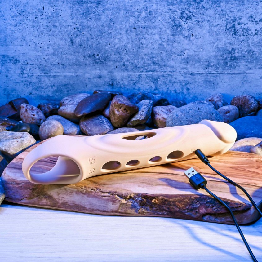 The Evolved Big Shaft Extender sits out on a table on top of a wooden slab. A charging cord is plugged into the extender to charge the vibrations. The charging port is right underneath the frenulum of the extender. | Kinkly Shop