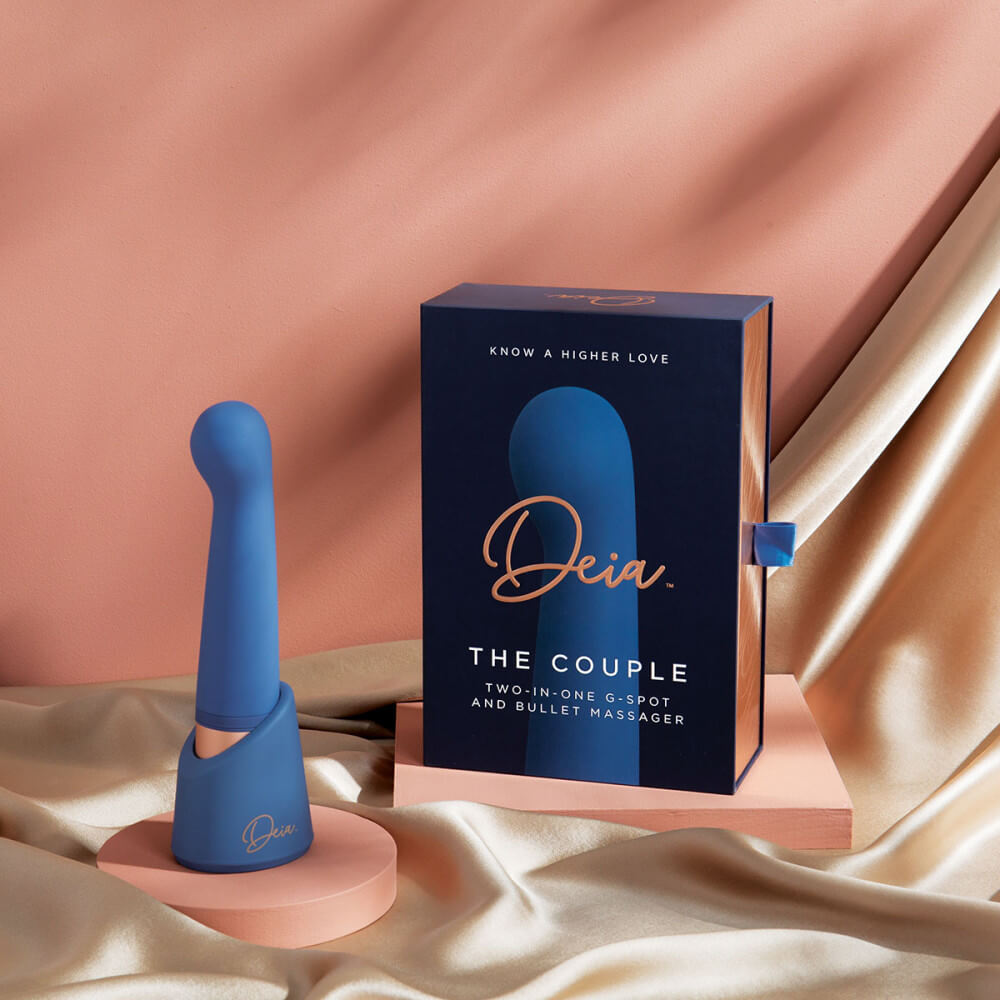 The Deia The Couple interchangeable tip vibrator is sitting upright in its charging/storage base right next to the packaging for the Deia The Couple interchangeable tip vibrator. The base is branded with the "Deia" logo and also color matches the vibrator itself. | Kinkly Shop