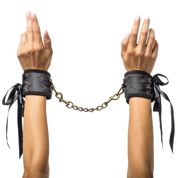 Two hands wear the cuffs included in the Kama Sutra Trust Me Erotic Playset. The cuffs are fastened with ribbons and have a satin-like finish. A gold chain with clips on both sides connects the cuffs to one another. | Kinkly Shop