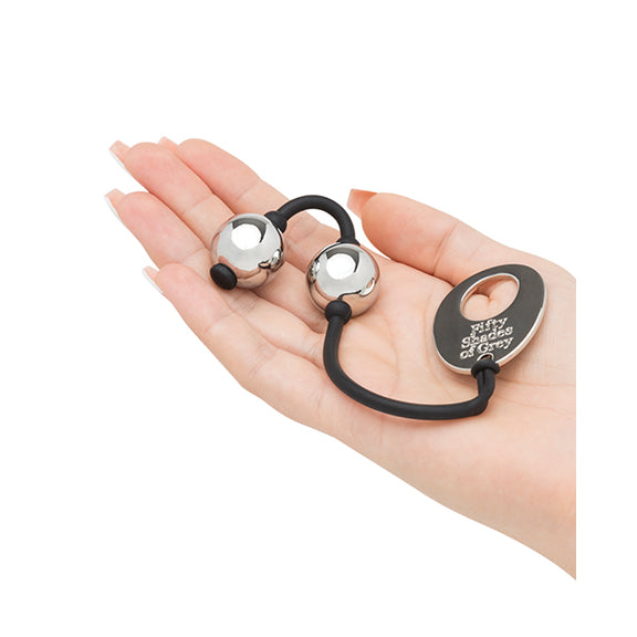 The Fifty Shades of Grey Inner Goddess Kegel Balls sitting inside of the flattened palm. The balls look particularly small and slim in the hands. The end of the kegel balls string has a large metal tag that has the "Fifty Shades of Grey" logo written on it. | Kinkly Shop