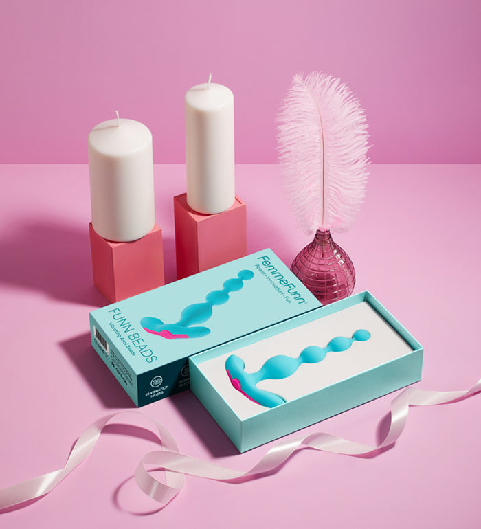 The FemmeFunn Funn Beads inside of its box. The box is opened, showcasing the toy inside. It's sitting in a pink display with pedestal pink candles and pink feathers and ribbon. | Kinkly Shop