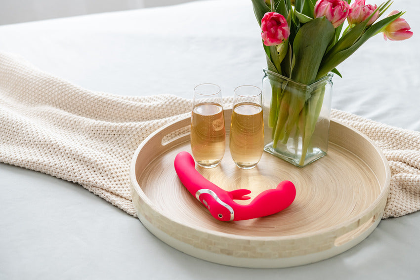 The Happy Rabbit Strapless Strap on Rabbit sitting out on top of a couch table shown next to two glasses of champagne with a vase of flowers. | Kinkly Shop