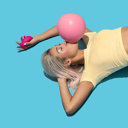A person lays on their back on a blue background. They are blowing a bubble in their mouth. They have the LELO SONA in their hand. The vibrator fits perfectly in the palm, looking like a comfortable object to grip. | Kinkly Shop