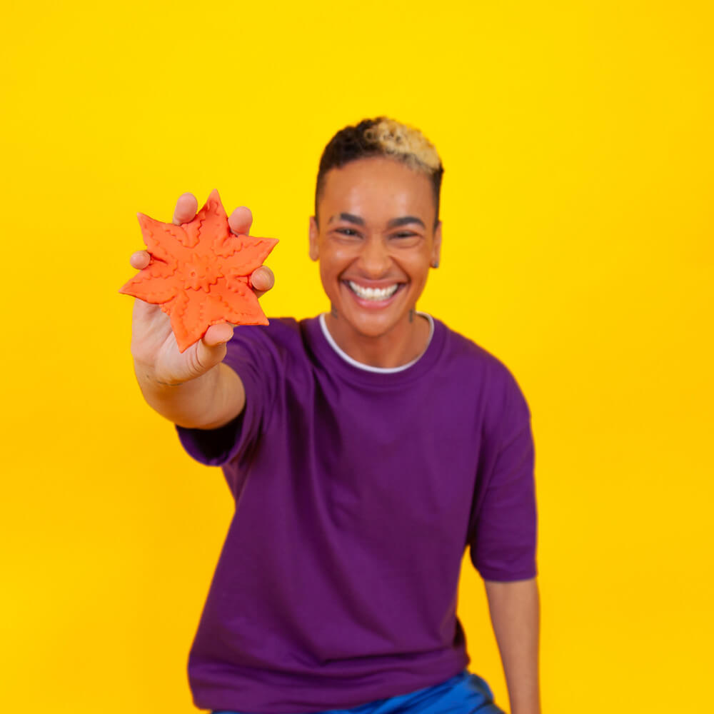 A smiling person against a yellow background is holding up a Coral Cute Little Fuckers Starsi to the camera. Their fingers comfortably fit in-between each one of the prongs of the design, and the Starsi is larger than their palm's size. | Kinkly Shop