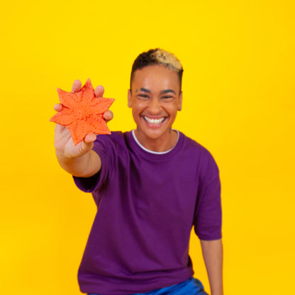 A smiling person against a yellow background is holding up a Coral Cute Little Fuckers Starsi to the camera. Their fingers comfortably fit in-between each one of the prongs of the design, and the Starsi is larger than their palm's size. | Kinkly Shop