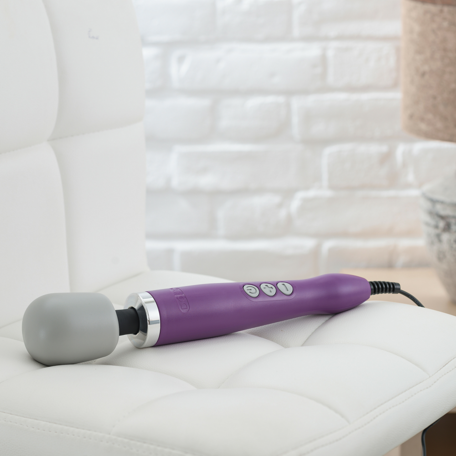 The Doxy Original Massager lays out on a chair. It's long, about as long as the chair, and the cord, which is unremovable, hangs off the end of the chair. | Kinkly Shop