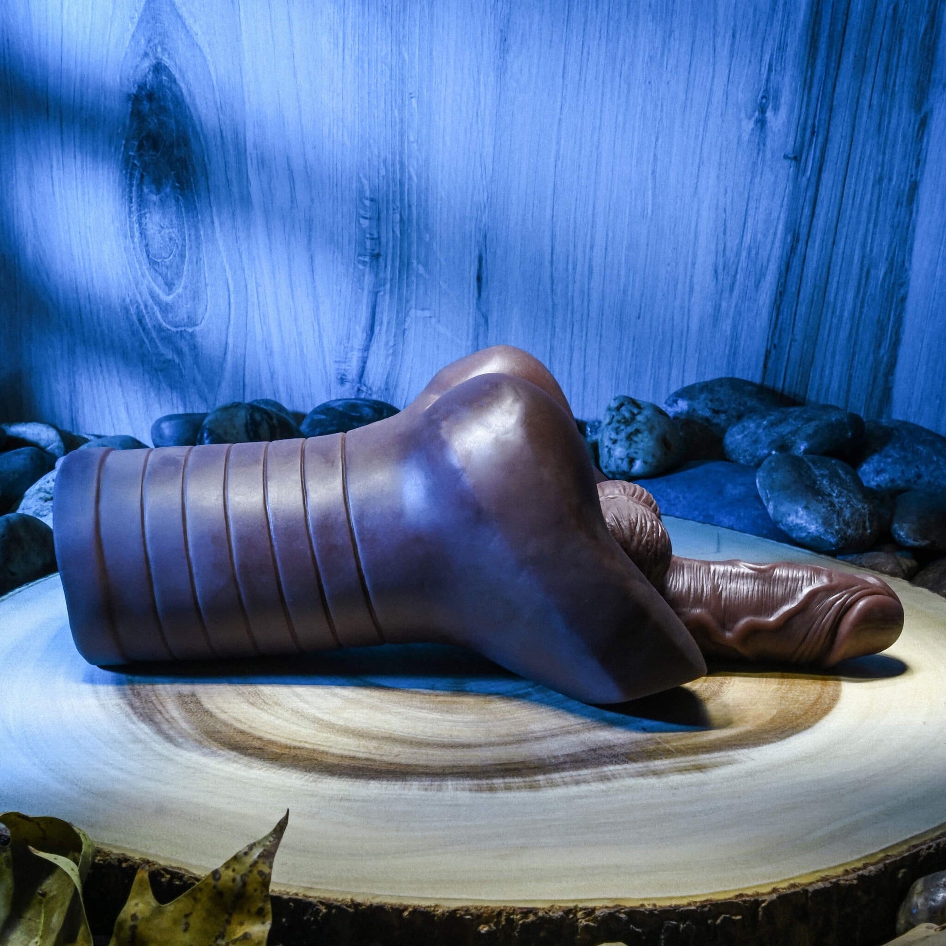 The Evolved Backdoor Bash in Espresso laying out on a slab of tree wood. Blue mood lighting really showcases the different textures of the Evolved Backdoor Bash including the ribbed structure that makes for easy handling and the very-textured penis shaft that protrudes from the stroker's entrance. | Kinkly Shop