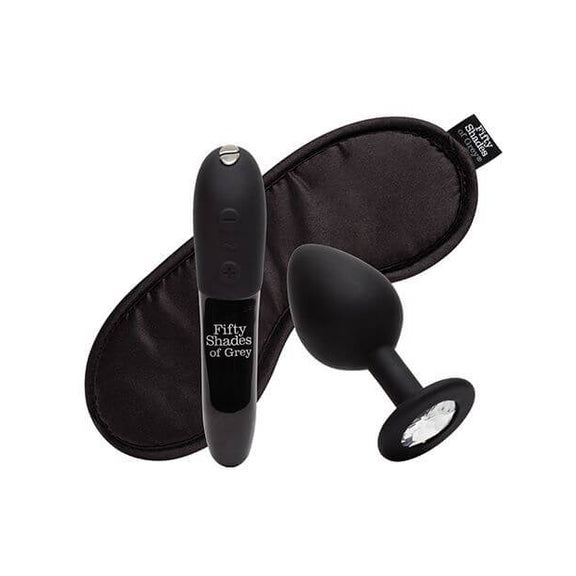 Everything included with the Fifty Shades of Grey Come to Bed Kit sits against a white background. There's the We-Vibe Tango X bullet vibrator in a black Fifty Shades of Grey design, a black butt plug with a circular, gem-shaped base, and a black, satin, branded Fifty Shades of Grey blindfold. | Kinkly Shop