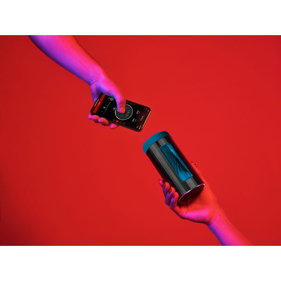 Two hands against a red background. One holds a LELO F1S V3 while another holds a cell phone, open to the LELO app to control the stroker. | Kinkly Shop