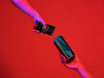 Two hands against a red background. One holds a LELO F1S V3 while another holds a cell phone, open to the LELO app to control the stroker. | Kinkly Shop