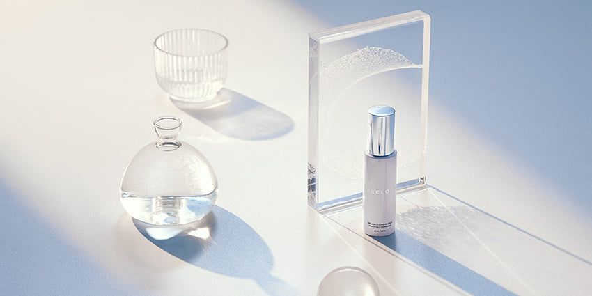 A bottle of the LELO Antibacterial (Toy) Cleaning Spray 2 fl oz sits out on a white surface with a strong sunbeam shining into the image from the corner. There are other white, see-through objects like glasses, cups, and blocks. All of the items cast soft, long shadows in the sunbeam. The toy cleaner bottle does as well. | Kinkly Shop