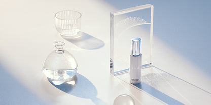 A bottle of the LELO Antibacterial (Toy) Cleaning Spray 2 fl oz sits out on a white surface with a strong sunbeam shining into the image from the corner. There are other white, see-through objects like glasses, cups, and blocks. All of the items cast soft, long shadows in the sunbeam. The toy cleaner bottle does as well. | Kinkly Shop