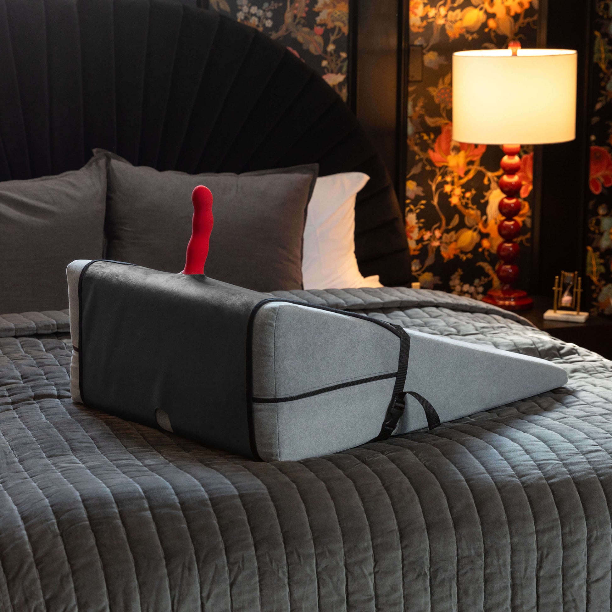 The Liberator Birdi Sex Toy Mount Add-On and the Ramp sitting out on top of a bed in a sultry-looking bedroom. A red dildo is protruding from the Ramp, held in place by the Birdi's mount system. | Kinkly Shop