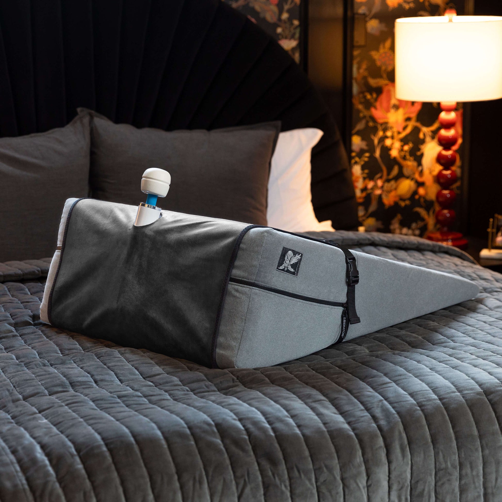 The Liberator Birdi Sex Toy Mount Add-On wrapped around a Ramp sitting on top of a bed in a sultry bedroom. A Vibratex Magic Wand wand massager is protruding from the top of the Ramp, snuggled into the toy mount function of the Birdi itself. The straps are wrapped snuggly around the width of the Ramp to keep the Birdi in place. | Kinkly Shop