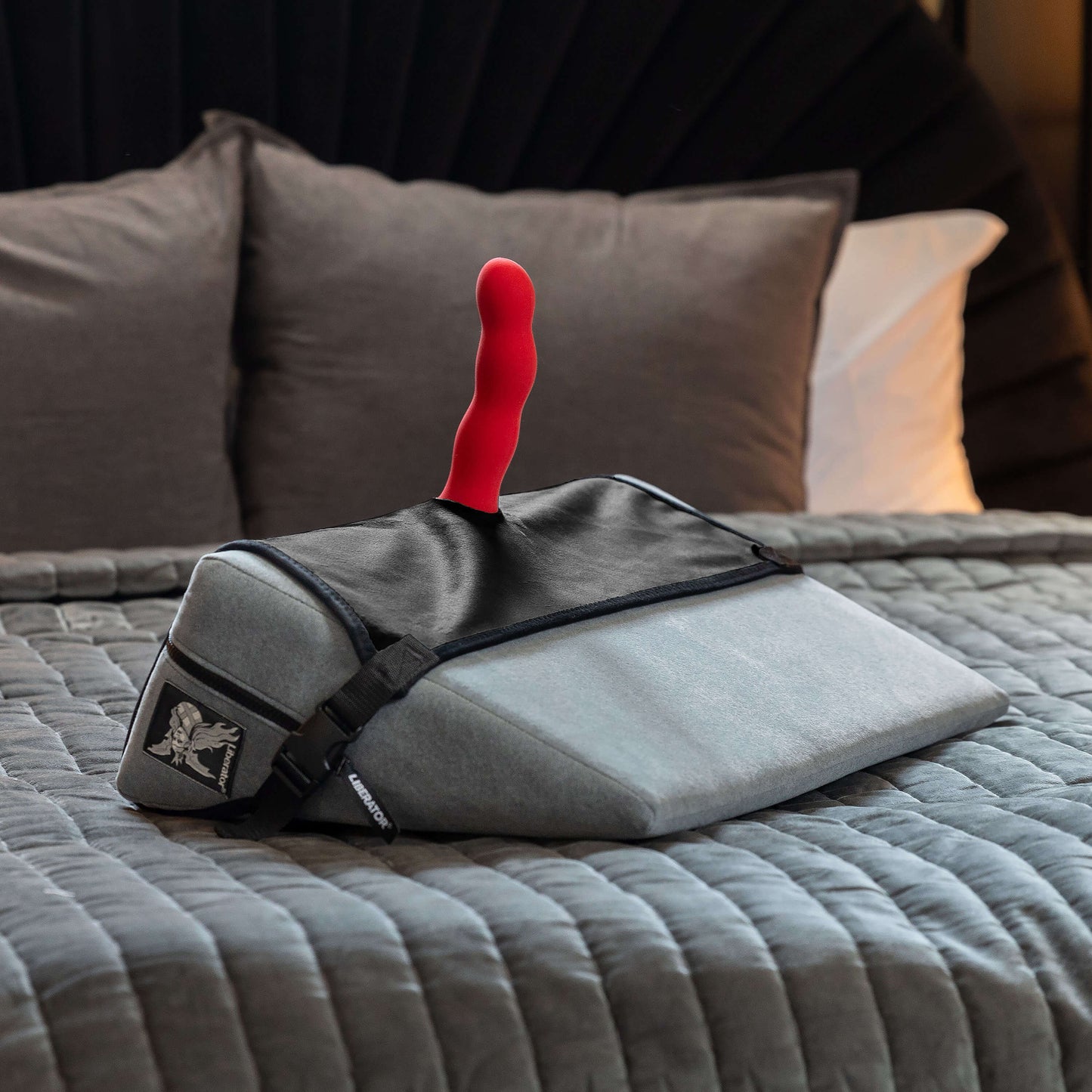 The Liberator Birdi Sex Toy Mount Add-On is wrapped around a Wedge, holding a red dildo protruding from the Wedge's surface. The Wedge and Liberator Birdi Sex Toy Mount Add-On sit on a bed in a sultry bedroom. | Kinkly Shop
