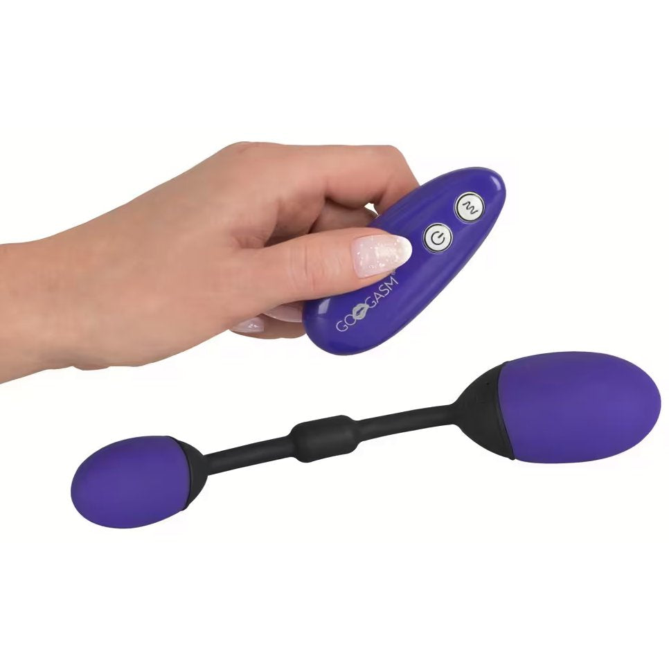A hand holds the GoGasm Vibrating Pussy & Ass Balls remote while the toy sits on the ground next to it. The remote has two buttons, and it looks like it'd be a bit too long to easily hide within a closed fist. | Kinkly Shop
