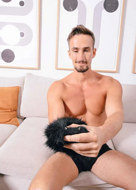 A person in their underwear sitting on a couch while wearing the Sportsheets Spiked Sensory Mitt on their hand. They are using their free hand to pull some of the faux fur aside to showcase the spikes built into the surface of the Sportsheets Spiked Sensory Mitt. | Kinkly Shop