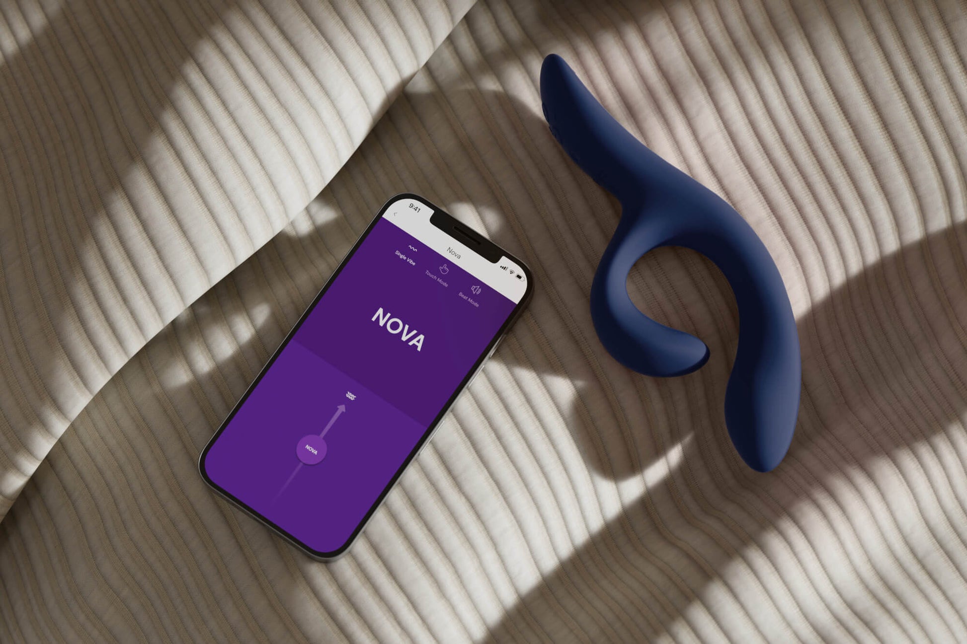 The We-Vibe Nova 2 lays flat on a bed. It's sitting out next to the app open on a cell phone. The cell phone app is showing the We-Vibe vibrator control app. | Kinkly Shop