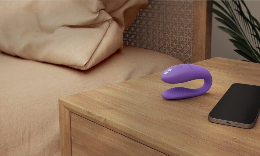 The We-Vibe Sync Go sits out near a cell phone on a bedside table. The We-Vibe Sync Go can be controlled by a free cell phone app. | Kinkly Shop