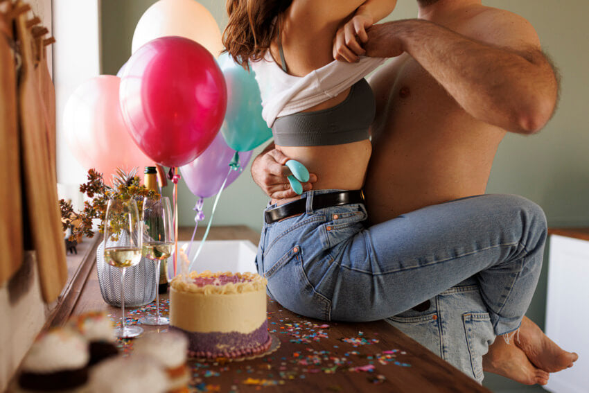 A couple is embracing. One of them is sitting on a table while their partner takes off the sitting person's shirt. One of the couple is holding an Aqua We-Vibe Sync Lite in their hand. The vibrator is surprisingly slender and about the size of one of the person's palms. | Kinkly Shop