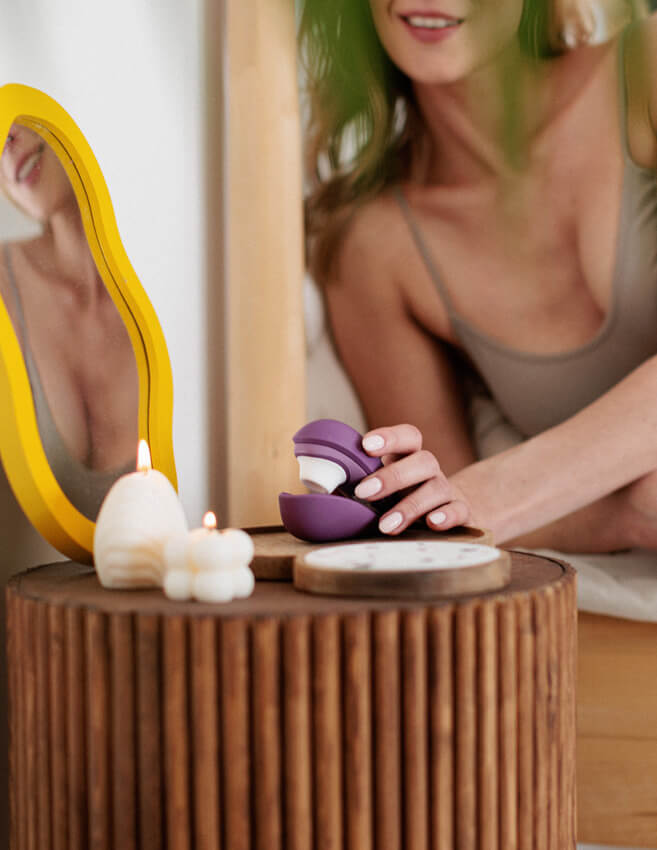 A person reaches onto their bedside table where a Womanizer Liberty 2 is sitting. The vibrator looks very slender, and it takes up less room on the side table than a nearby candle. | Kinkly Shop