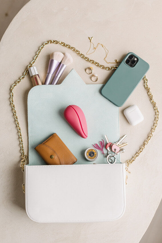 The Womanizer Liberty 2 with its lid attached is laying out next to an open purse. Other purse items are scattered around it. The vibrator is shorter and smaller than make-up brushes or the cell phone, and it's about twice the length of an Earbuds case. | Kinkly Shop