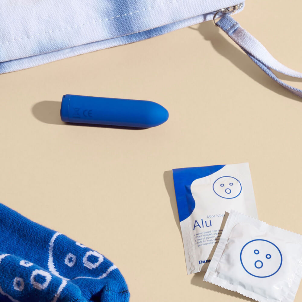The Dame Zee micro bullet vibrator set out on a beige background near other Dame items. The image clearly shows packets of Dame Alu lube as well. The Dame Zee micro bullet vibrator is about the same size as these sample size lube packets and slightly longer than the standard square size of a condom. | Kinkly Shop