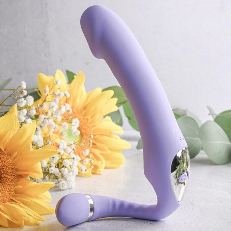 The Gender X Orgasmic Orchid sits upright in front of a display of yellow flowers and green leaves. The smaller "tail" end has been curled around in a semi-circle to act as a "stand" to get the shaft upright to point towards the ceiling. | Kinkly Shop