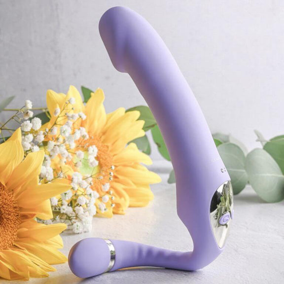 The Gender X Orgasmic Orchid sits upright in front of a display of yellow flowers and green leaves. The smaller "tail" end has been curled around in a semi-circle to act as a "stand" to get the shaft upright to point towards the ceiling. | Kinkly Shop
