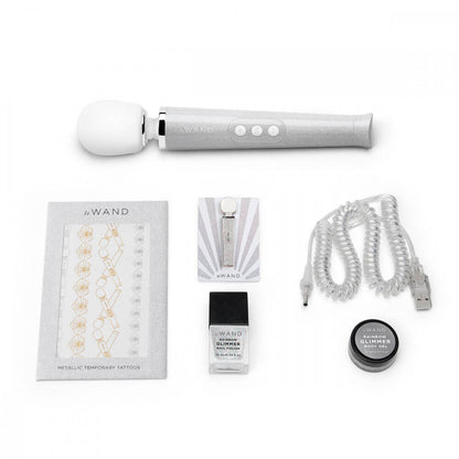 The White Le Wand All That Glimmers Wand Massager laid out next to everything that comes with the petite wand massager. That includes the color-coded charging cable, a glittery body gel, a matching enamel pin, a glittery nail polish, and a set of color-matching metallic temporary tattoos. | Kinkly Shop