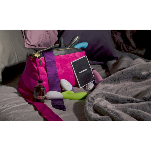 A Liberator Tallulah Locking Toy Bag sitting open on a plush bed. All of the items that were inside of the Liberator Tallulah Locking Toy Bag are tossed next to the bag. The bag is unzipped. Multiple dildos are poking out the top. Multiple toys are scattered around its bottom. A bottle of lube is sitting next to the bag as well. | Kinkly Shop