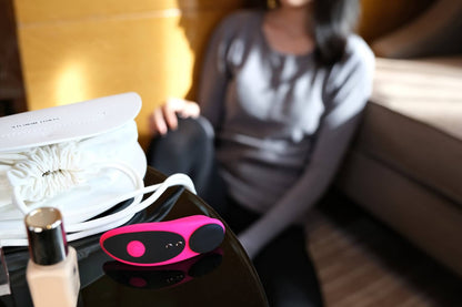 Lovense Ferri sits on a table in the foreground while someone in a long-sleeve tee is leaning up against a couch in the background. The Ferri appears to be about the size of a small bottle of nail polish or concealer. | Kinkly Shop