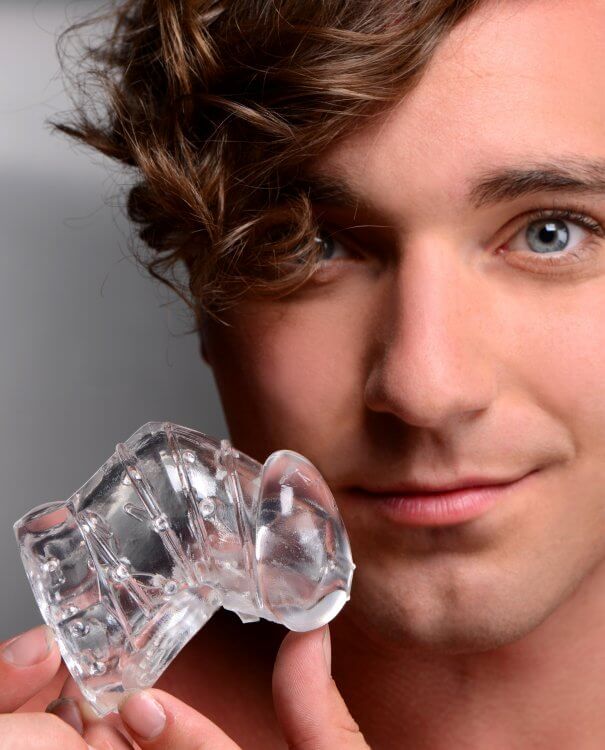 A person with blue eyes smiles into the camera while holding the Master Series Detained 2.0 Restrictive Chastity Cage. The built-in downward angle of the cage is very evident from this angle, and it looks surprisingly short. | Kinkly Shop
