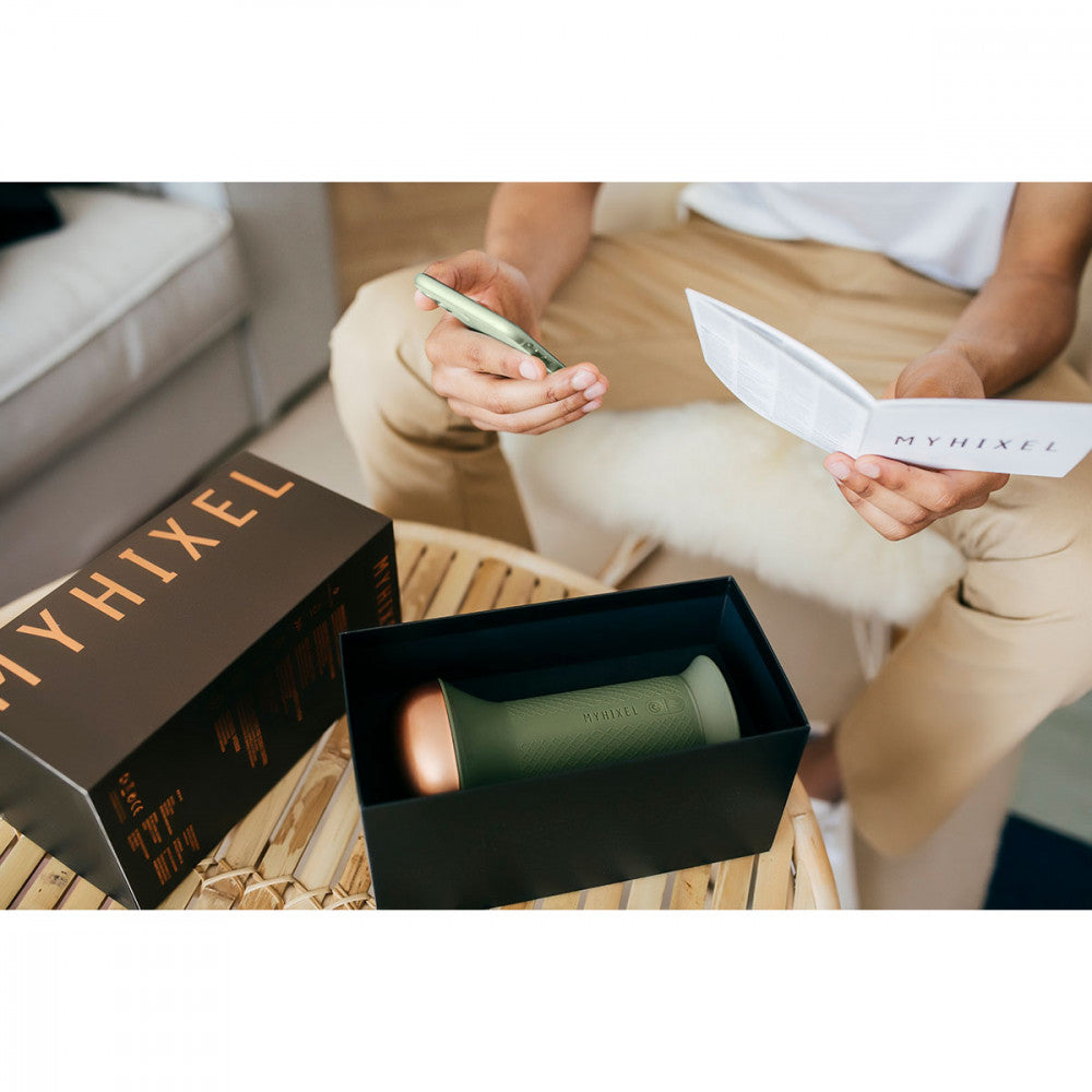 A person is reading the MyHixel TR manual in one hand and installing the MyHixel Play app on their cell phone in the other hand. On the table in front of them, the MyHixel TR premature ejaculation sex toy lays inside of its open box. | Kinkly Shop