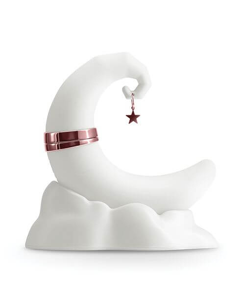 The NS Novelties Firefly Dream Glow up against a white background in daylight. It's extremely white, and it almost blends into the background. The toy is crescent moon shaped with a star charm hanging off the tip. It sits upright without the help of a hand because it's resting in its charging base that looks like a pillowy cloud. | Kinkly Shop