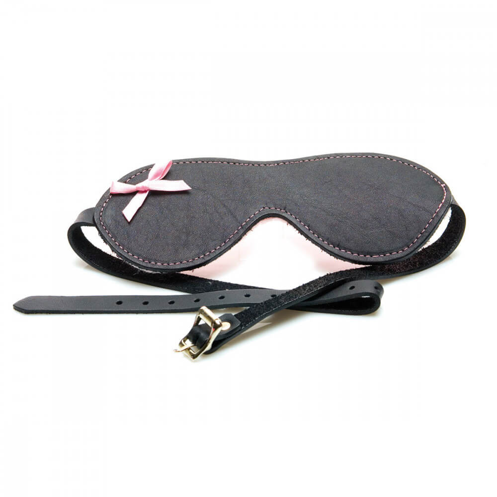 The Sex Kitten Leather Buckle Blindfold laying out on a white surface. The front of the Sex Kitten Leather Buckle Blindfold is smooth, black leather with a small, pink bow in the upper left corner of the blindfold. This image shows the buckle adjustment system for getting a good fit. | Kinkly Shop