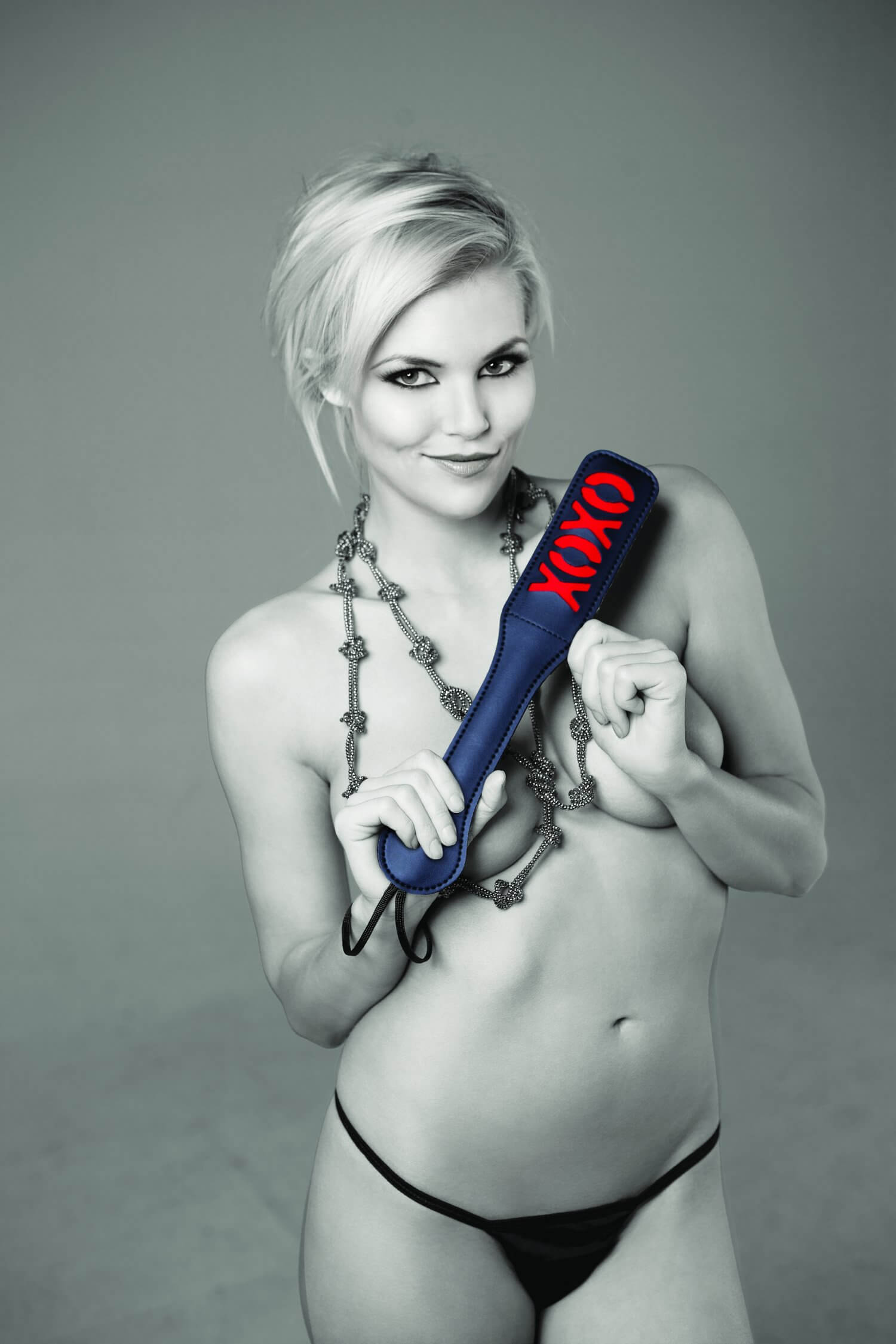 A person smiling while they hold the Sportsheets Black Vinyl Paddle, XOXO slapper in front of their body. | Kinkly Shop