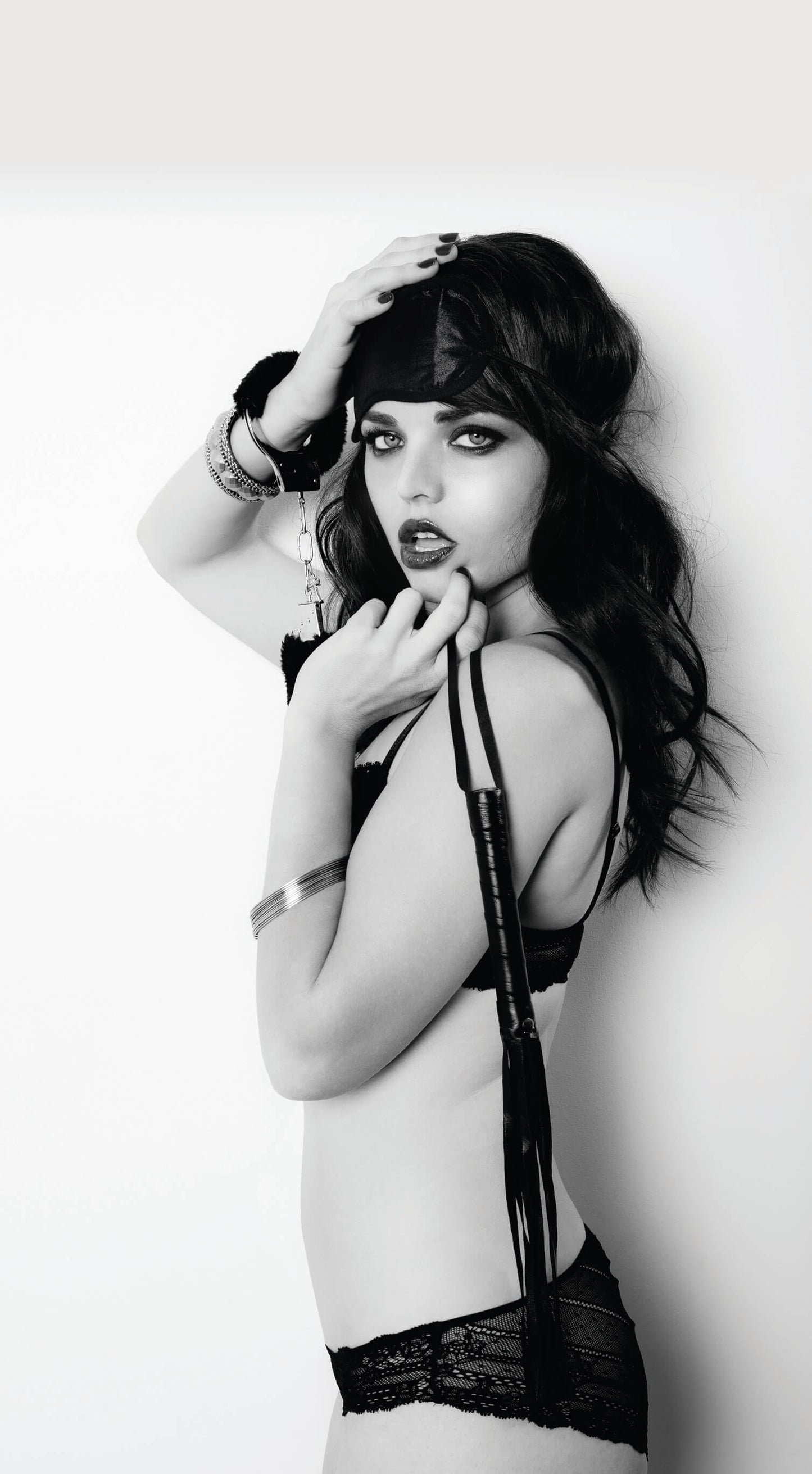 A person wearing the Intro to S&M kit. They are wearing the blindfold on their forehead, have the faux fur cuff wrapped around one wrist, and are dangling the flogger by its wrist strap. They are dressed in lingerie. | Kinkly Shop