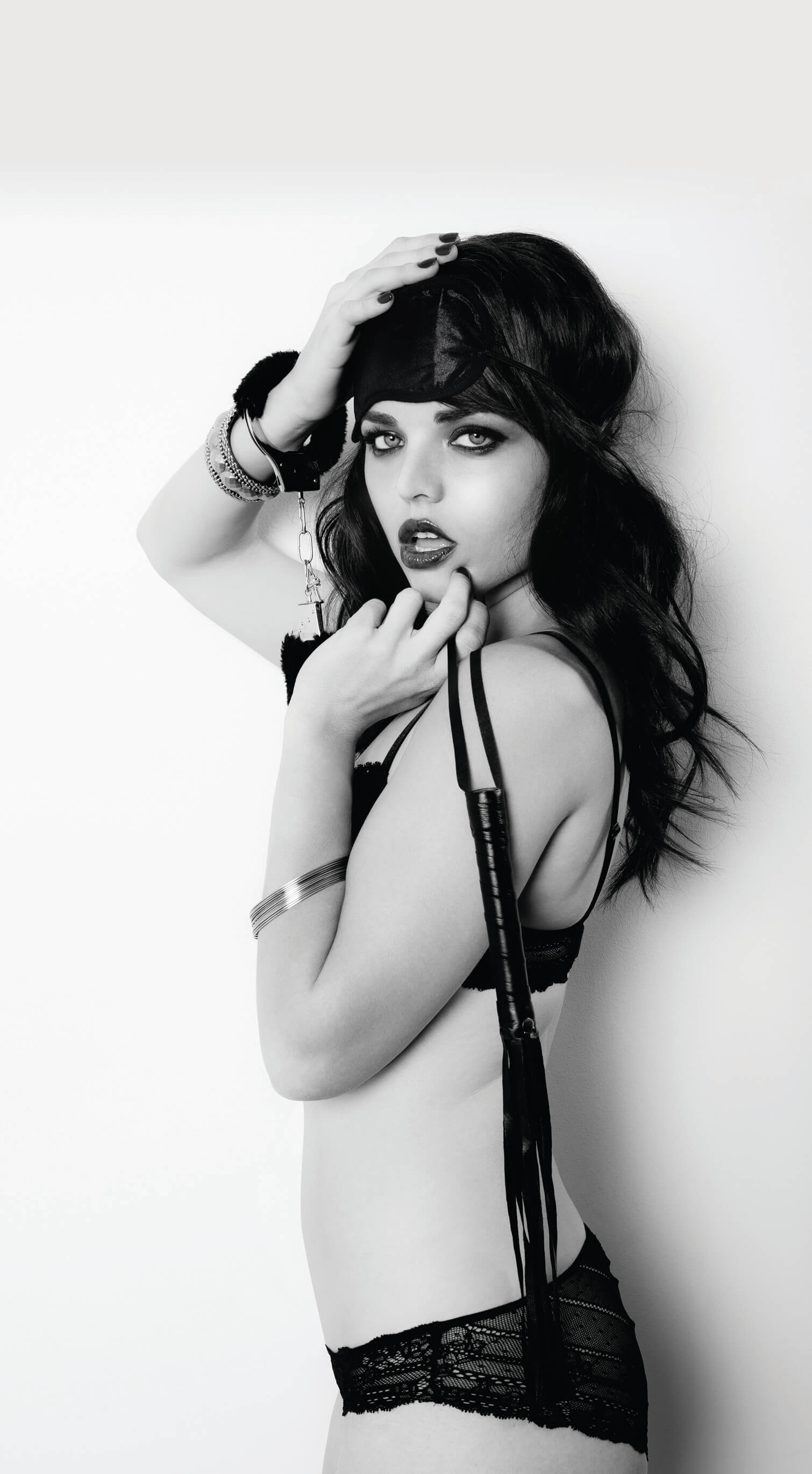 A person wearing the Intro to S&M kit. They are wearing the blindfold on their forehead, have the faux fur cuff wrapped around one wrist, and are dangling the flogger by its wrist strap. They are dressed in lingerie. | Kinkly Shop