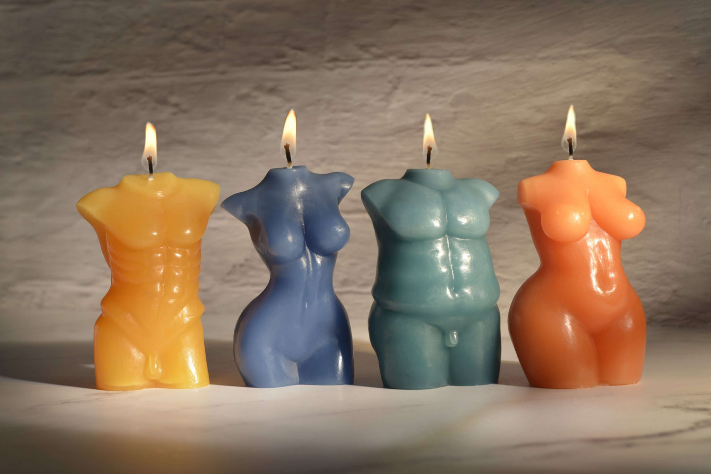 All of the four Sportsheets Torso Form Wax Play Candles sitting next to one another on top of a marbled surface. There's dark, moody lighting, and all of the candles are lit. This showcases all of the sculpted details of each candle. | Kinkly Shop