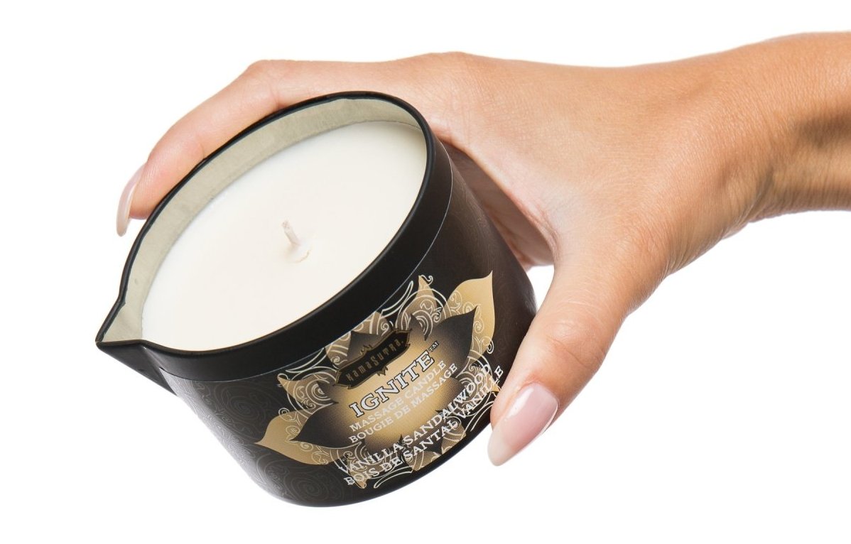The Kama Sutra Ignite Massage Candle: A hand is depicted holding a one-wick candle made of white wax in a black jar. The side of the jar has a spigot for pouring the wax onto a person's skin as it melts.